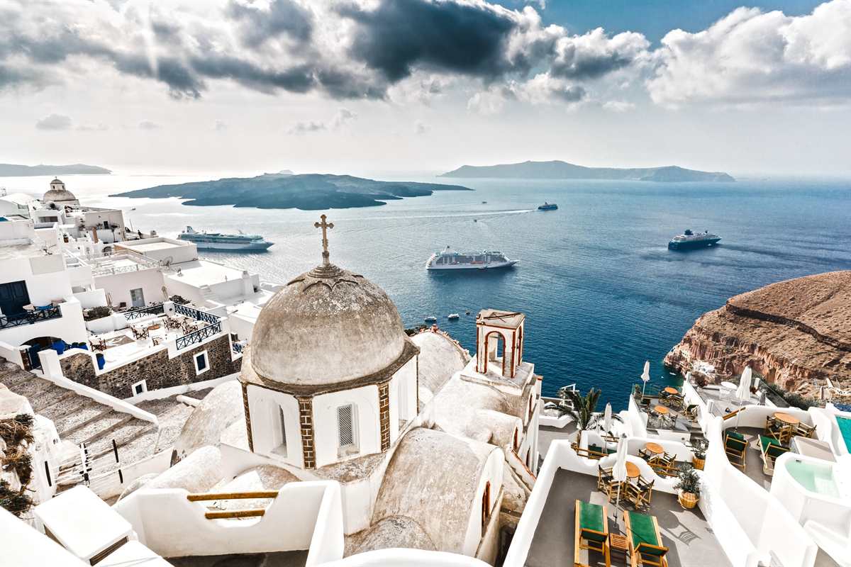 Santorini shore excursion: a white church with a grey stone dome and a cross above the caldera at Fira, with three cruise ships at anchor on deep blue water and the dark volcanic islands beyond under a cloudy sky