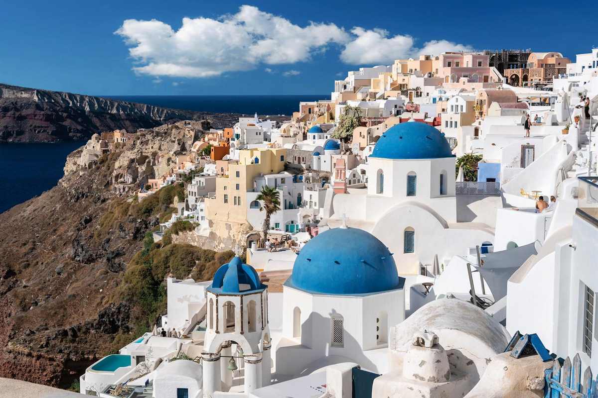 Santorini shore excursion: two bright blue church domes and a blue-topped white bell tower of Oia above a cliff of white and pastel houses, with the dark blue caldera sea and a brown cliff beyond under a deep blue sky with white clouds