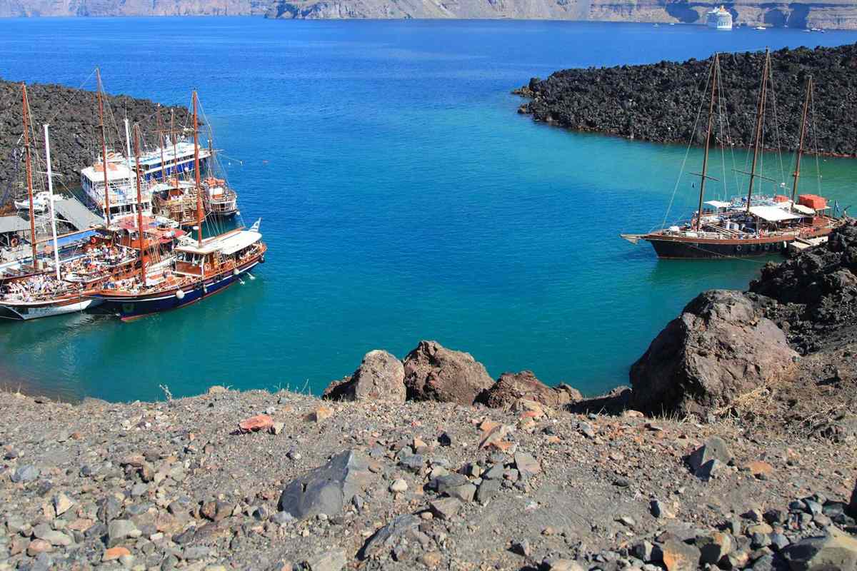 Santorini volcano hot springs tour: wooden tour boats moored in the turquoise bay at Nea Kameni, with black lava rocks in front and the caldera cliffs of Santorini across the water