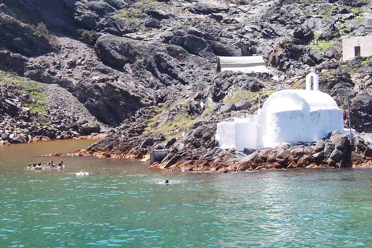 Santorini volcano hot springs tour: swimmers in the orange-brown warm water of the Palea Kameni hot springs bay, beside a small white chapel on the dark volcanic rocks
