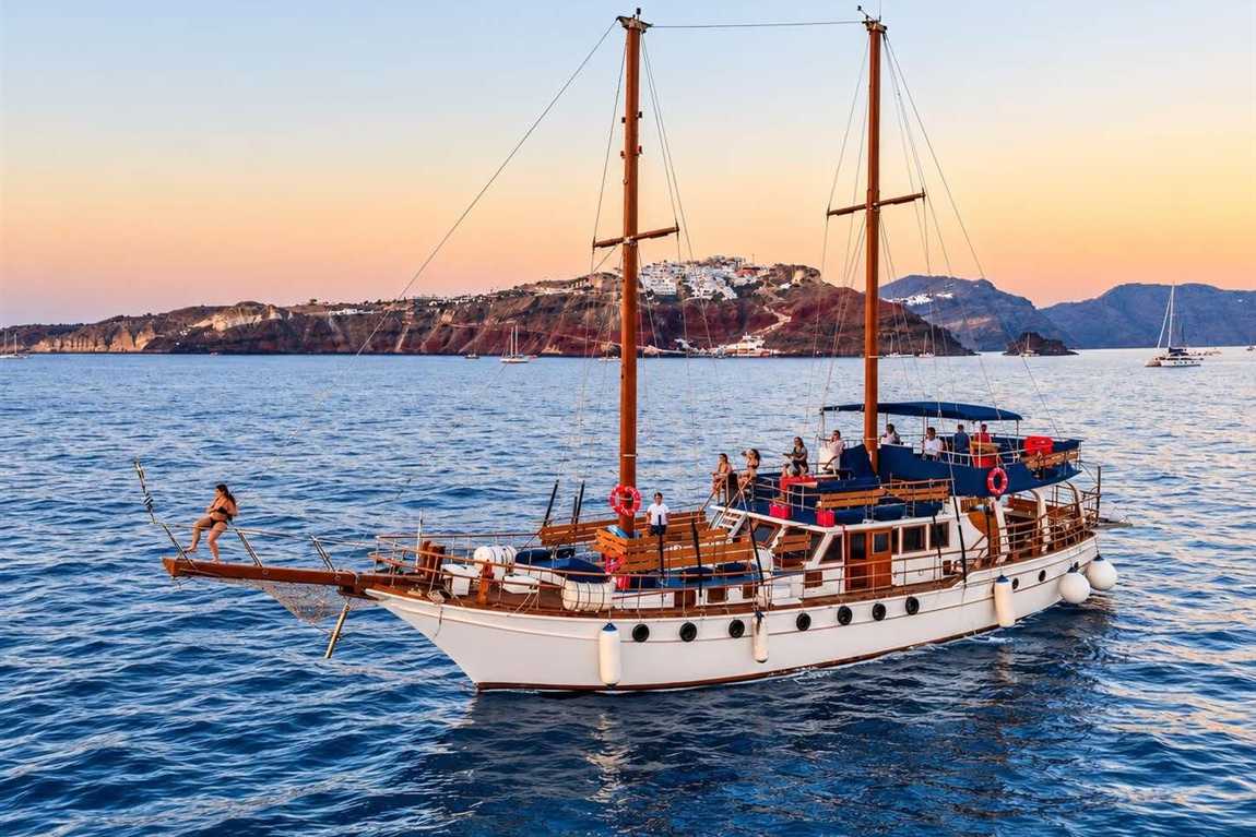 Santorini volcano sunset cruise: a two-masted white and wooden cruise boat with blue deck cushions sailing on the deep blue caldera at dusk, with the red-brown caldera cliffs and white village of Santorini and a hazy orange sky behind
