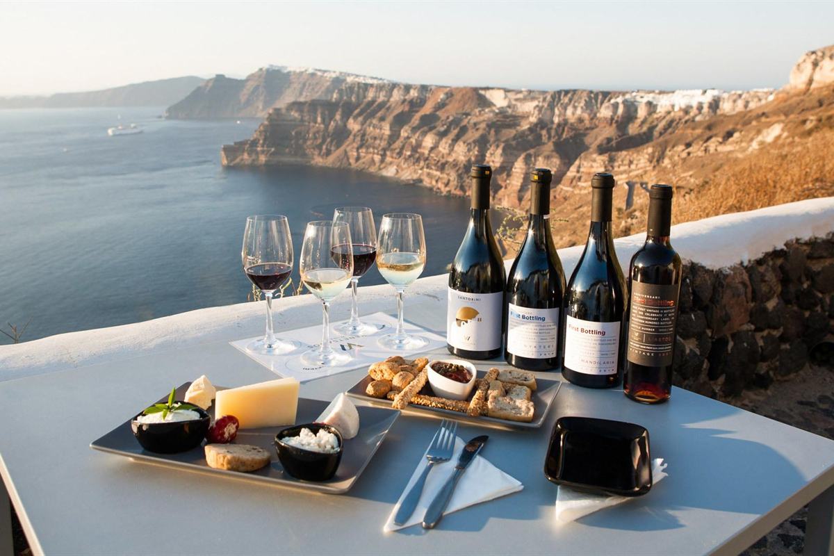 Santorini wine tour with sunset in Oia: a tasting table on a white terrace wall above the caldera, set with four glasses of red and white wine, four wine bottles and plates of cheese, dips and breadsticks, with the layered orange cliffs and blue water of the caldera behind in warm evening light