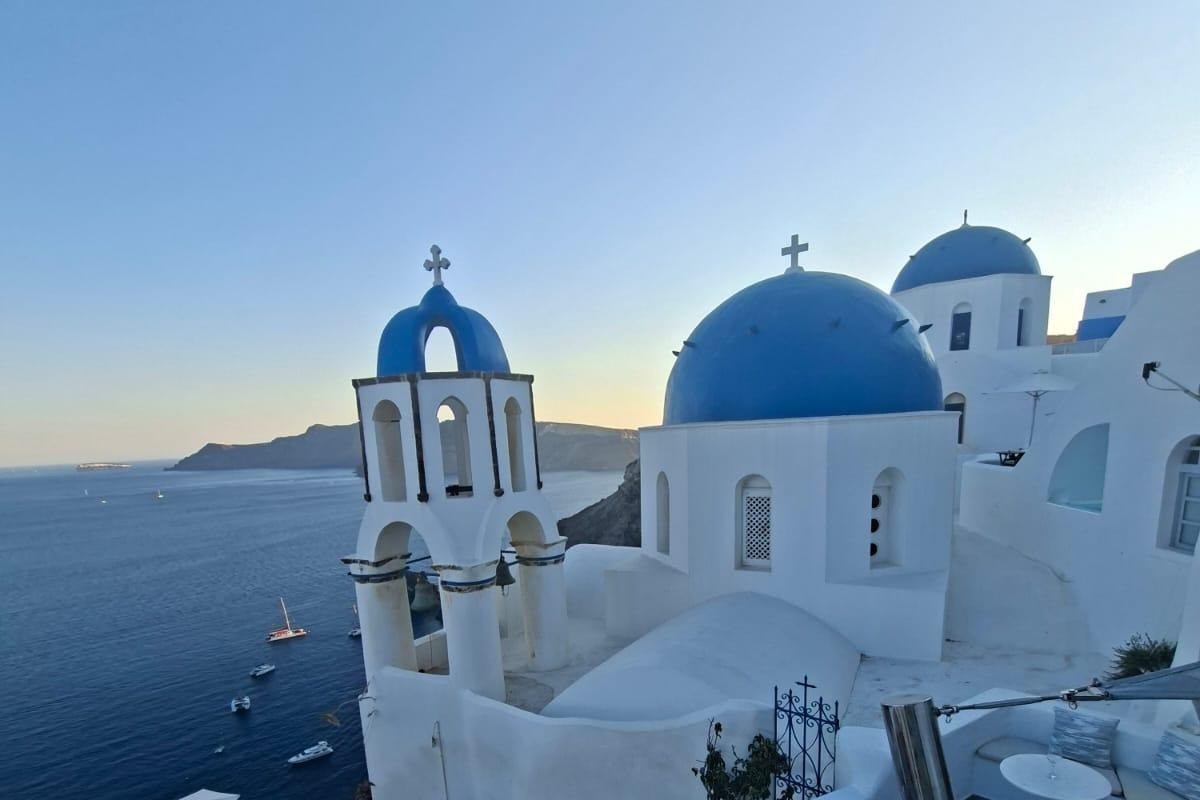 Santorini wine tour with sunset in Oia: a white bell tower with a blue arch beside a blue-domed church in Oia above the deep blue caldera, with small boats on the water and a soft orange glow on the horizon at dusk