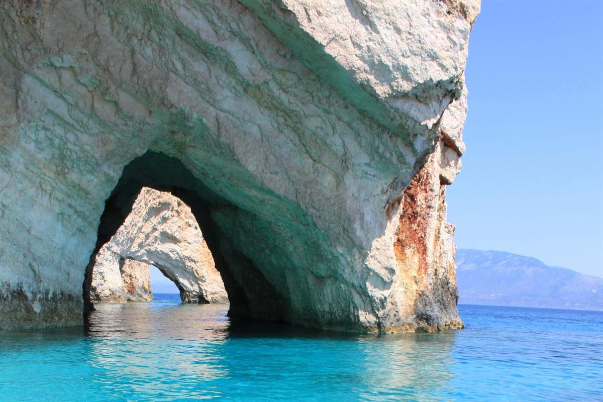 Shipwreck beach tour Zakynthos: pale limestone sea arches of the Blue Caves rising from clear turquoise water, with a second arch behind the first and distant hills under a blue sky