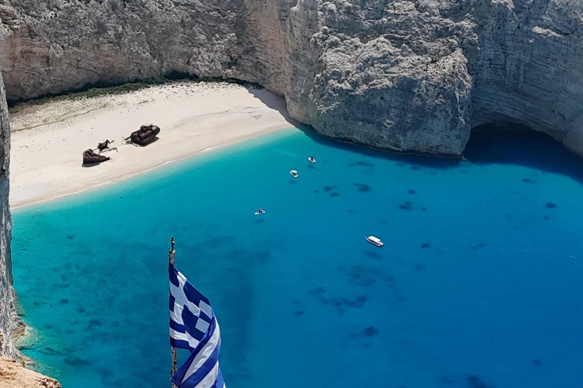 Shipwreck beach tour Zakynthos: view from the cliff top down into Navagio bay, with the rusted shipwreck lying on the white sand below tall pale limestone cliffs, small boats floating on the turquoise water and a Greek flag in the foreground