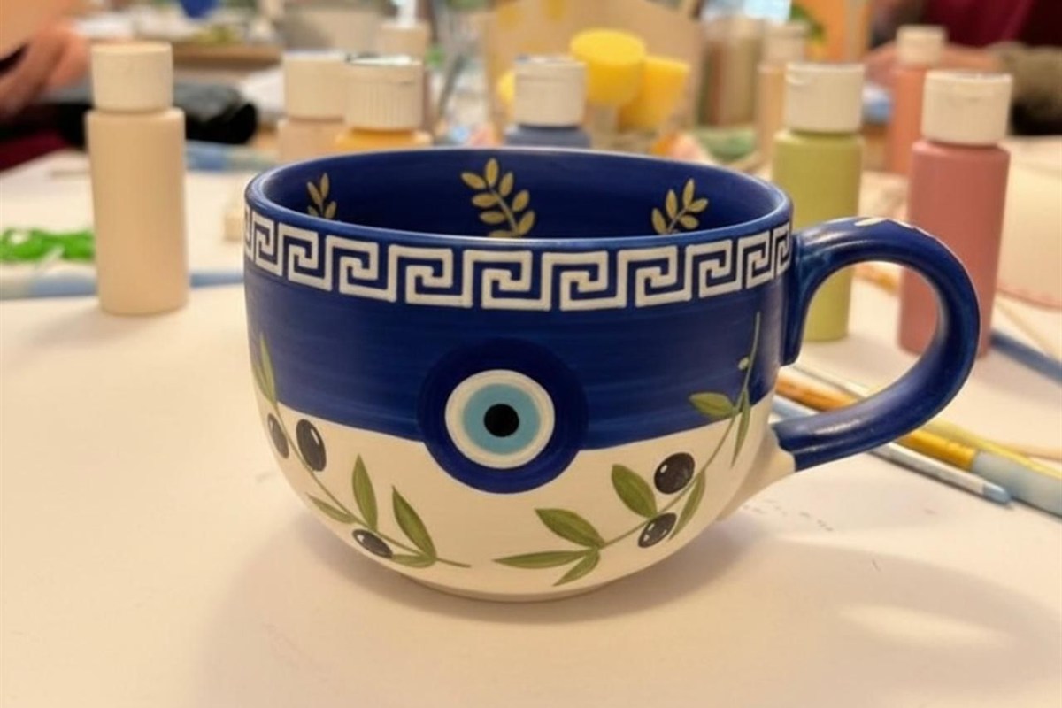 Sip and paint Thessaloniki: a ceramic mug painted dark blue and white with a white Greek key border, a blue evil eye and olive branches with black olives, standing on a white table in front of bottles of acrylic paint, brushes and yellow sponges