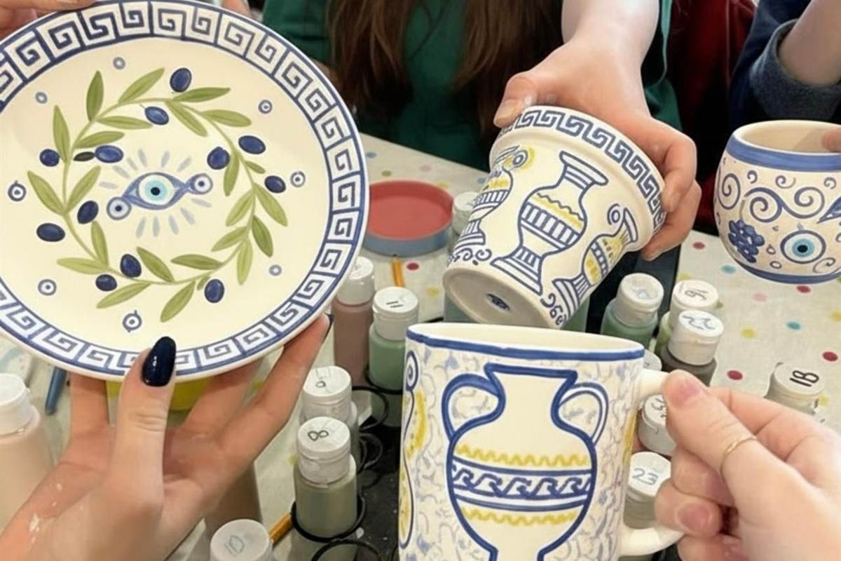 Sip and paint Thessaloniki: hands holding a plate painted with a blue Greek key border, green olive branches and an evil eye, beside a mug with blue amphora vases and a second painted mug, above a table of numbered acrylic paint bottles