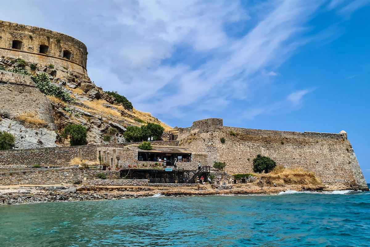 Spinalonga day trip from Agios Nikolaos: the round Venetian bastion and long stone sea wall of the Spinalonga fortress above a small waterside taverna and rocky shore, with turquoise water in front and a blue sky with thin white clouds