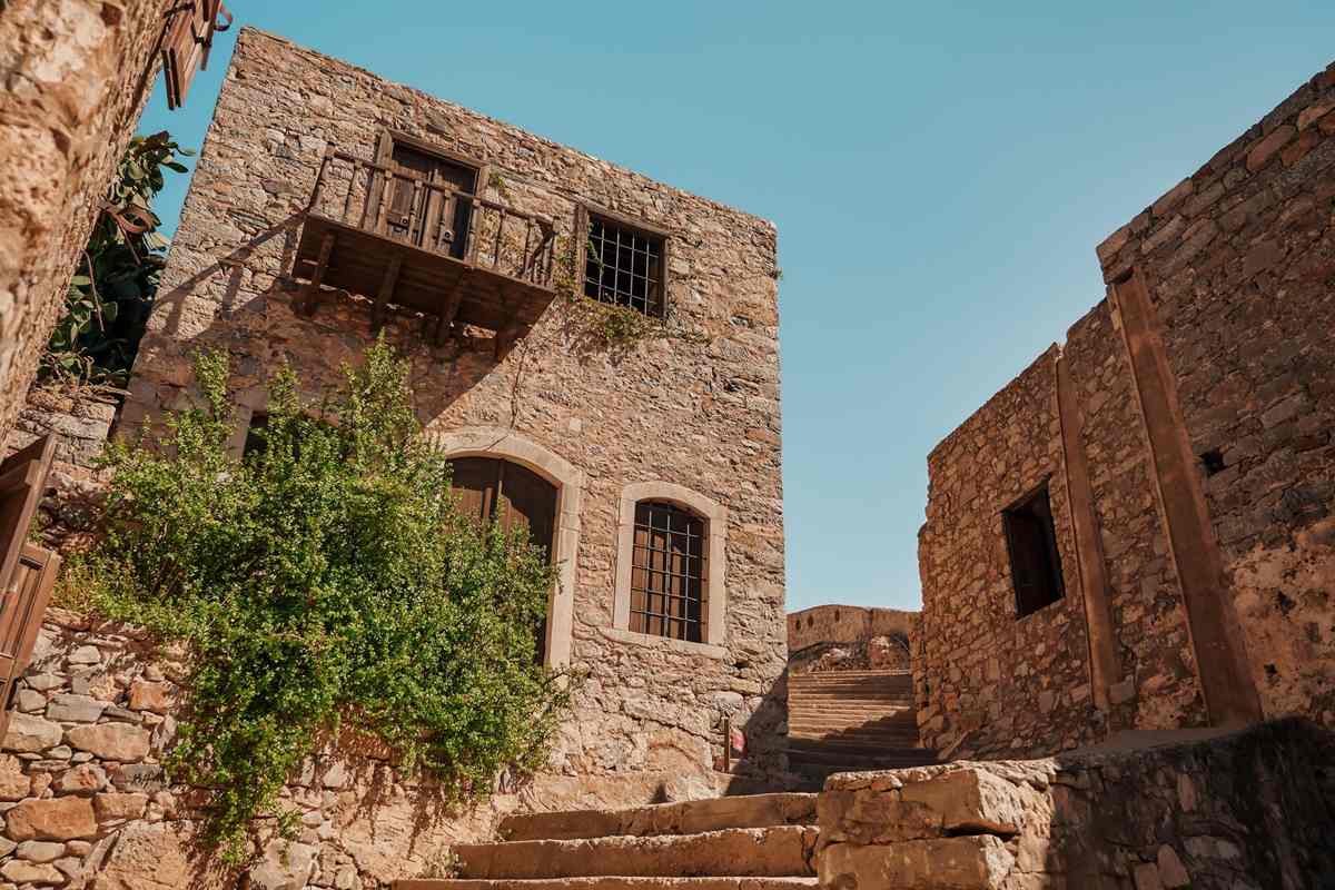 Spinalonga day trip from Agios Nikolaos: a narrow stone lane inside the Spinalonga fortress, with a two-storey stone house that has a wooden balcony and barred arched windows, green shrubs beside it and worn stone steps climbing under a clear blue sky