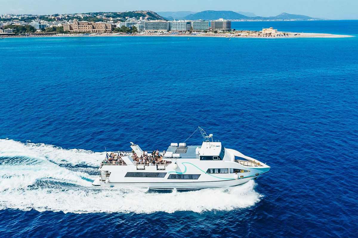 Symi day trip from Rhodes: a white passenger boat speeds across deep blue water leaving a white wake, with the hotels and beach of Rhodes town behind it