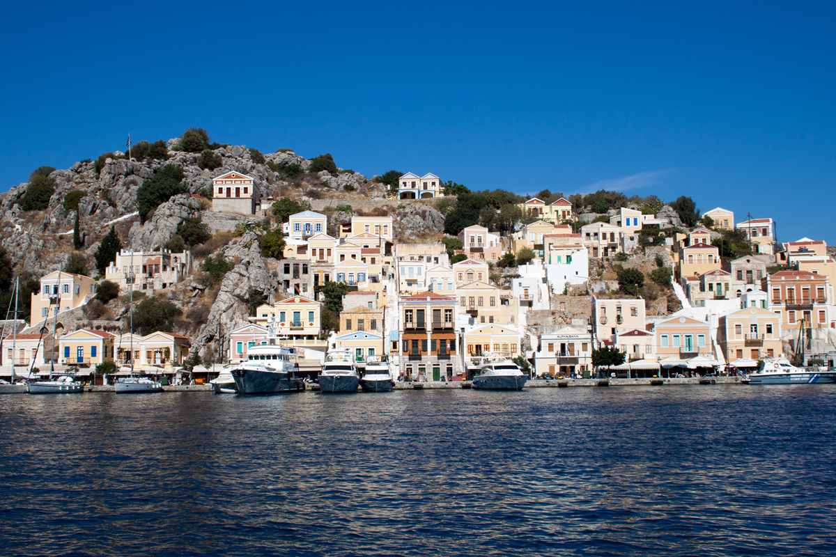 Symi day trip from Rhodes: pastel neoclassical houses climbing the rocky hill above Gialos harbour, with yachts moored along the quay under a clear blue sky
