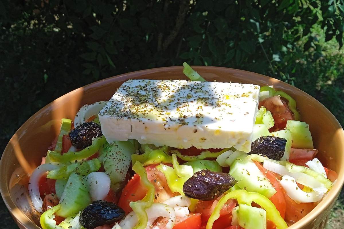 Thessaloniki cooking class: a Greek village salad in a brown ceramic bowl, topped with a thick slab of white feta sprinkled with dried oregano, over red tomato wedges, green pepper rings, cucumber, white onion and dark olives, with dark green garden leaves behind