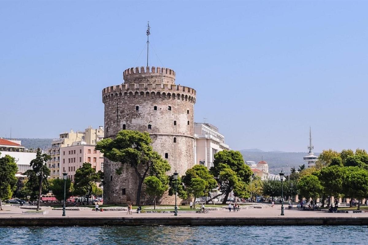 Thessaloniki cooking class: the round White Tower with its crenellated stone top and a flag on a tall pole, seen from the water across the stone quay, with green pine trees, street lamps and city buildings under a clear blue sky