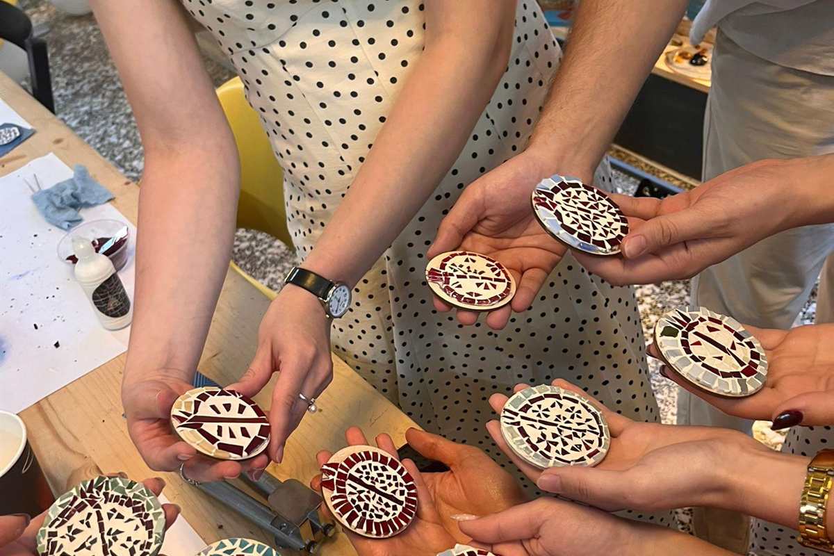Thessaloniki mosaic magnet: hands holding round handmade mosaic pieces made of small red, cream and grey tiles above a wooden workshop table