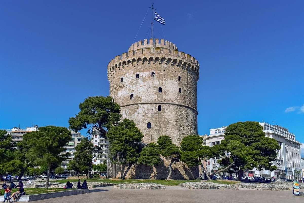 Thessaloniki mosaic magnet: the round White Tower of Thessaloniki with its stone battlements and a Greek flag on top, standing behind green pine trees on a paved waterfront square under a deep blue sky
