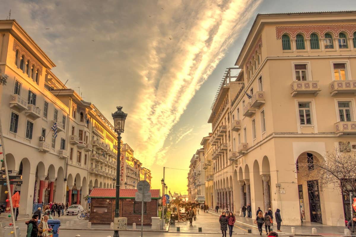 Thessaloniki underworld night walk: a street between cream-coloured arcaded buildings with balconies at sunset, a black lamp post in the middle and a long golden cloud streak across the evening sky, with walkers in the distance