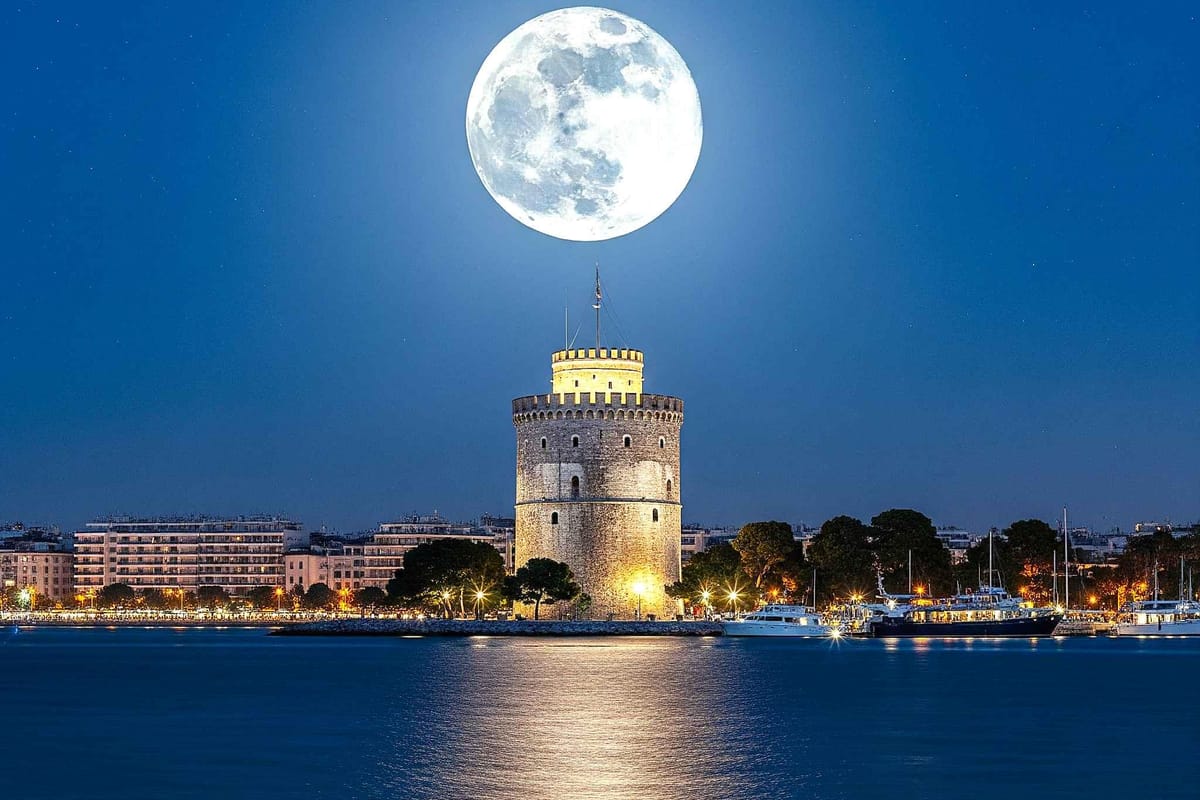 Thessaloniki underworld night walk: the round White Tower with its lit stone battlements on the waterfront under a huge full moon, with city lights and moored boats reflected in the dark blue sea