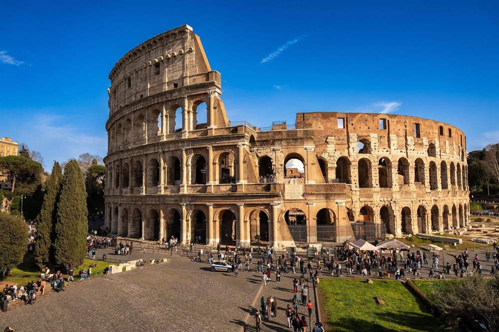 Tours of Italy feature the Colosseum in Rome, a large ancient stone amphitheatre with rows of arches under a clear blue sky, with visitors walking on the paved square in front.
