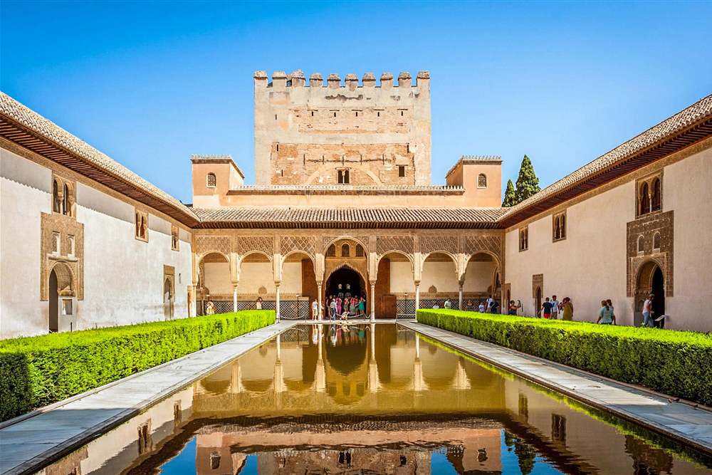 Tours of Spain: a long reflecting pool in a palace courtyard with trimmed green hedges, arched galleries and a crenellated stone tower beyond, under a clear blue sky