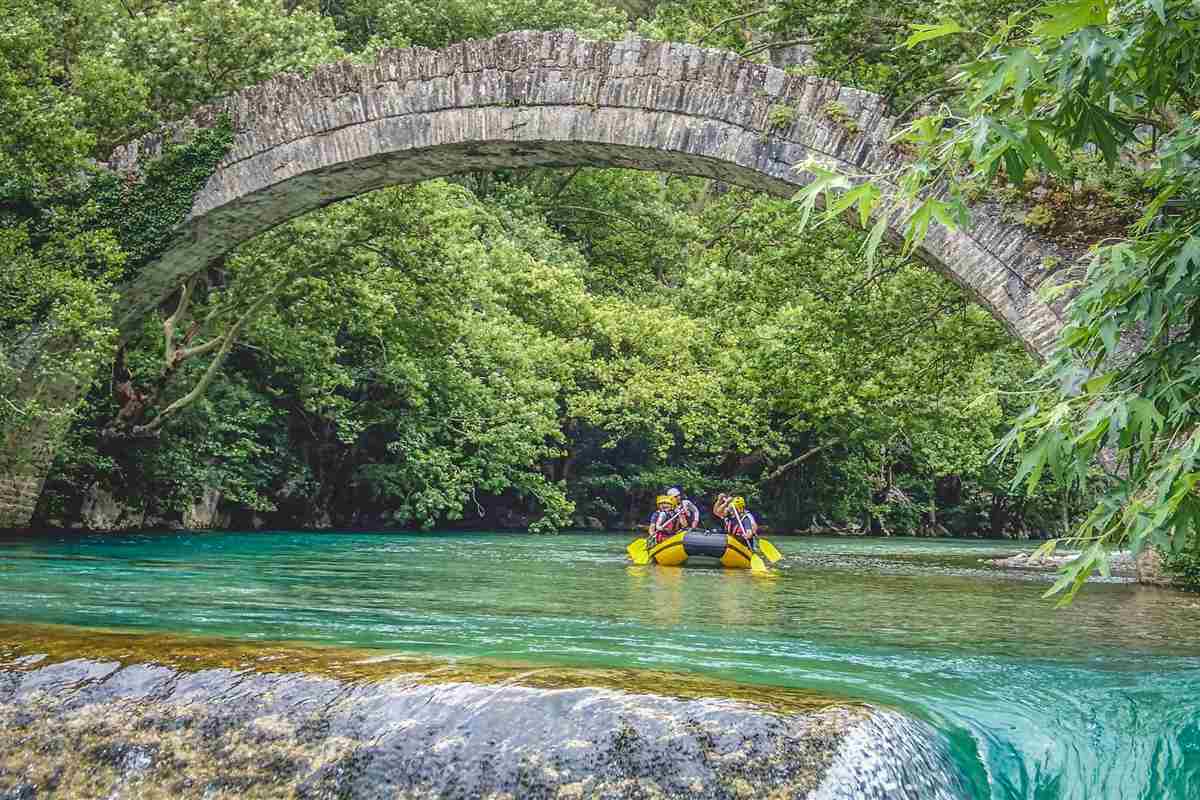 Voidomatis rafting: a yellow raft with a small crew in helmets floating on turquoise river water beneath the tall single arch of an old stone bridge, framed by dense green plane trees