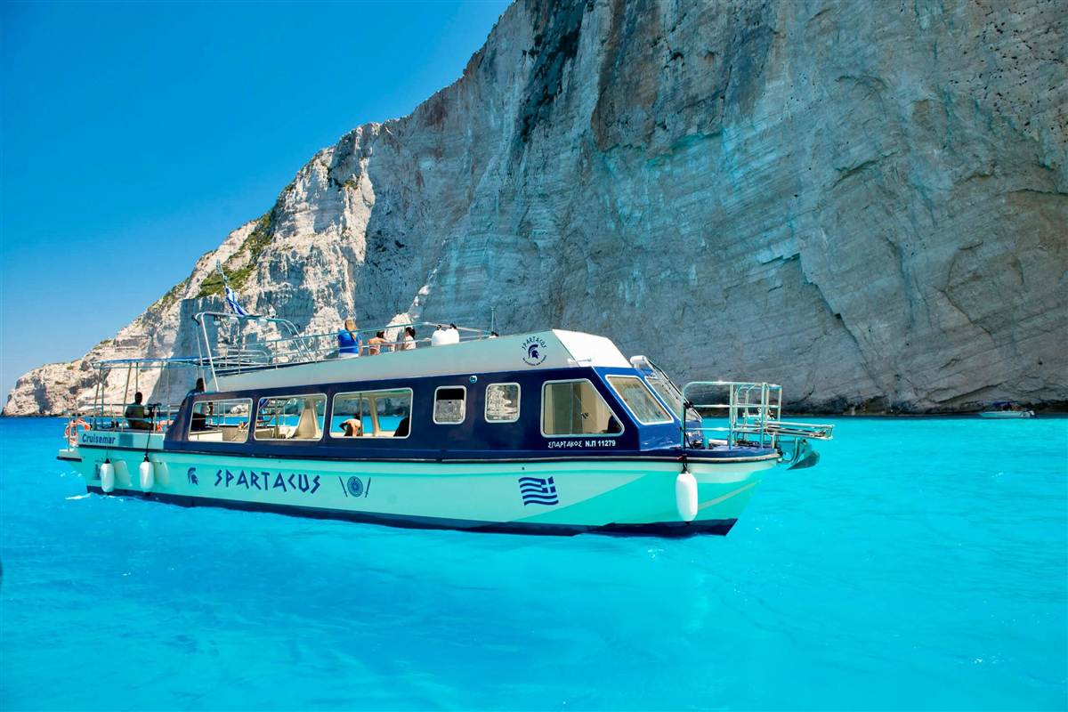 Zakynthos glass bottom boat: a white and navy two-deck glass-bottom boat with large side windows and a Greek flag floating on bright turquoise water beneath a tall pale limestone cliff under a clear blue sky