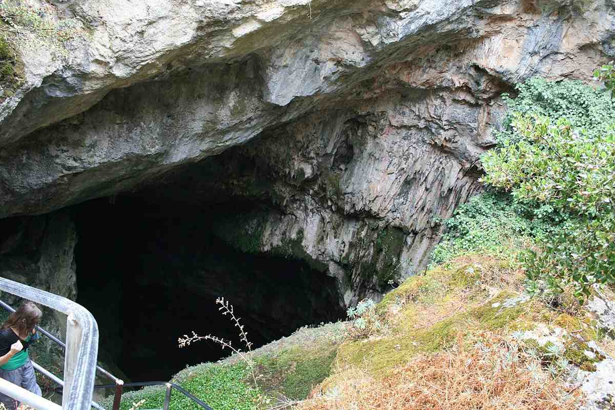 Zeus Cave Lasithi Plateau tour: the wide mouth of a cave in a grey limestone cliff, with stone steps and a metal handrail at the lower left, green shrubs on the right and dry grass in front